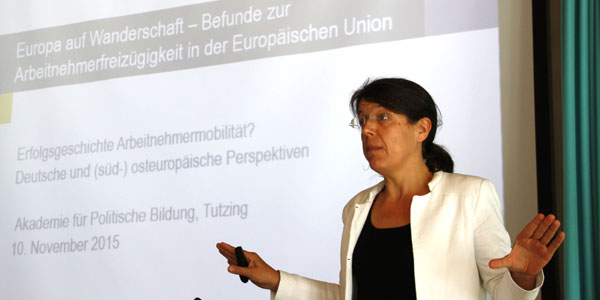 Carola Burkert im Hörsaal der Akademie für Politische Bildung auf einem Podium und hält einen Vortrag. Links von ihr  ist eine Powerpoint-Präsentation zu sehen 