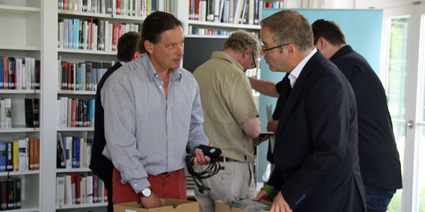 Steffen Elsner und Armin Heil stehen sich an einem Tisch gegenüber und reden. Steffen Elsner hält in der rechten Hand ein Kabel und Armin Heil hält einen Karton. Im Hintergrund stehen drei weitere Männer neben Bücherregalen. 
