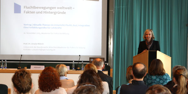 Ursula Münch steht in einem Hörsaal der Akademie für Politische Bildung am Rednerpult und begrüßt die Gäste, die vor ihr sitzen. Links neben ihr sind Tische und eine Leinwand, auf der eine PowerPoint-Präsentation zu sehen ist