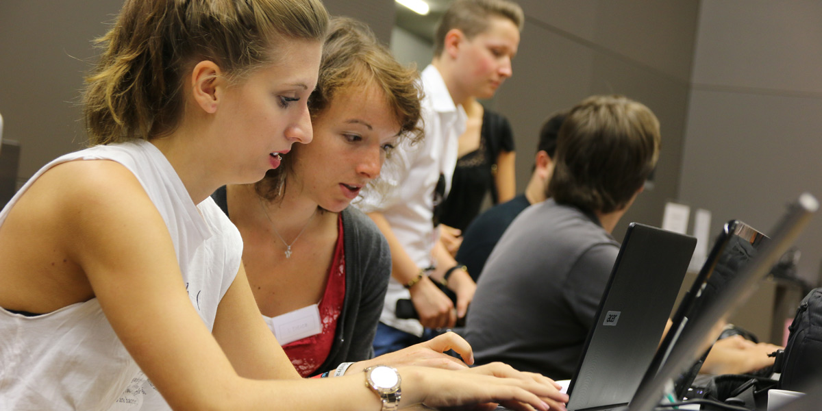 Zwei junge Journalistinnen arbeiten tippen etwas in ihren Laptop. Neben ihnen sitzen weitere junge Journalisten, die zusammen Gruppenarbeit betreiben