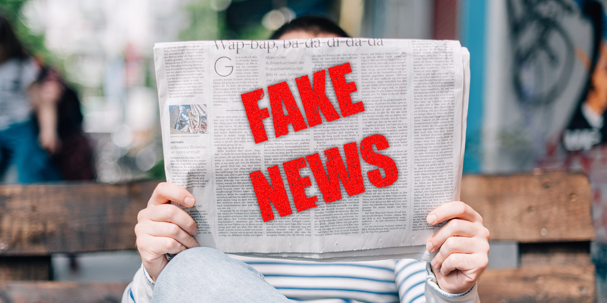 Eine Person sitzt auf einer Holzbank und hält eine Zeitung vor das Gesicht. Auf der Zeitung steht in roter Schrift "FAKE NEWS"