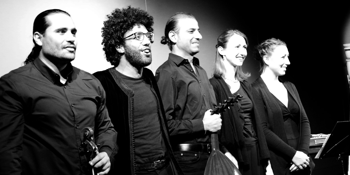 Schwarz-Weiß-Fotografie: Mohcine Ramdan, Ehab Abou Fakhir, Lisa Schöttl, Christine Horter und ein weiterer Mann stehen auf dem Podium des Hörsaals der Akademie für Politische Bildung und lächeln in die Kamera. Ehab Abou Fakhir hält eine Oud in der Hand