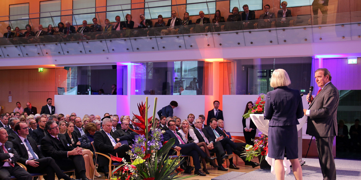 erhard Schröder und Ursula Münch stehen auf einem Podium in der IHK und halten jeweils ein Mikrofon. Vor ihnen sowie an den Seiten über ihnen, sitzen Personen und schauen interessiert nach vorne