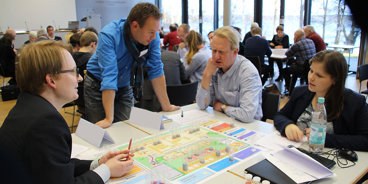 Im Vordergrund sind vier Personen an einem Tisch und spielen ein Planspiel. Im Hintergrund sitzen weitere Personen an Tischen, die auch Planspiele durchführen