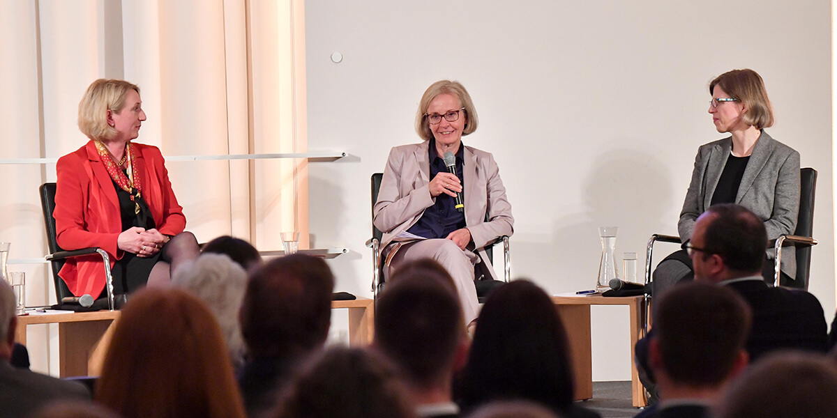 Diane Ahrens, Ursula Münch und Anne Margarian sitzen auf einem Podium, vor Publikum. Neben ihnen ist jeweils ein kleiner Tisch mit einer Wasserkaraffe und einem Glas. Diane Ahrens und Anne Margarian haben zudem ein Mikrofon neben sich liegen. Ursula Münch hält ihres in der Hand