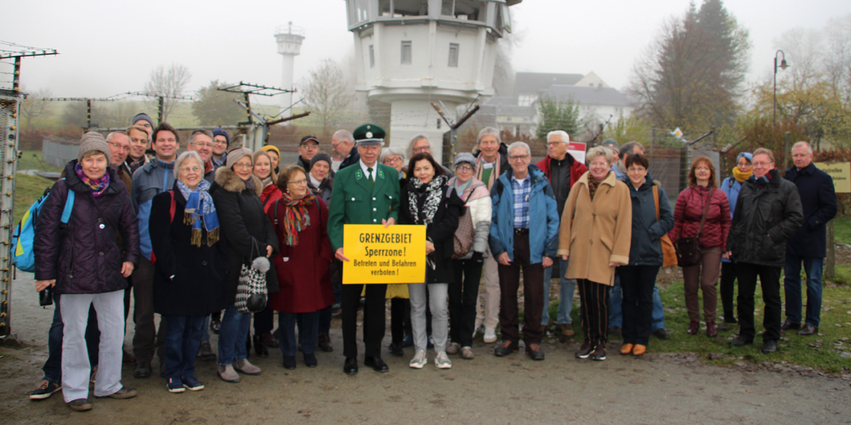 Personen stehen mit einem ehemaligen Grenzpolizisten an dem früheren Grenzgebiet. Der ehemalige Grenzpolizist hält ein gelbes Schild hoch, auf dem draufsteht, dass das Betreten der Sperrzone verboten ist