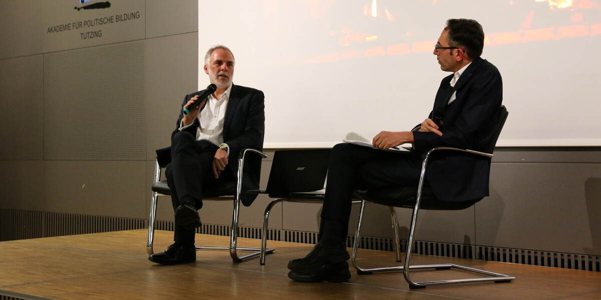 Alexander Kmentt und Hakan Akbulut sitzen auf dem Podium des Hörsaals der Akademie für Politische Bildung Tutzing. Beide halten ein Mikrofon in der Hand