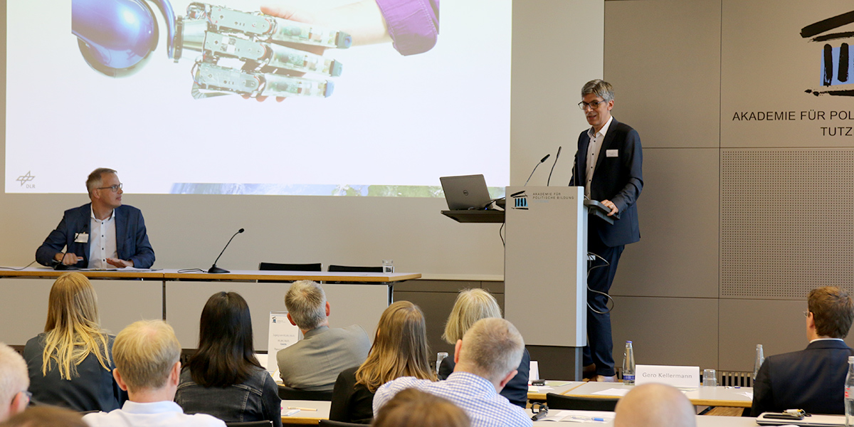 Gero Kellermann sitzt im Hörsaal der Akademie für Politische Bildung Tutzing an einem Tisch. Vor ihm sitzt das Publikum und rechts von ihm steht Alin Albu-Schäffer am Rednerpult und hält einen Vortrag
