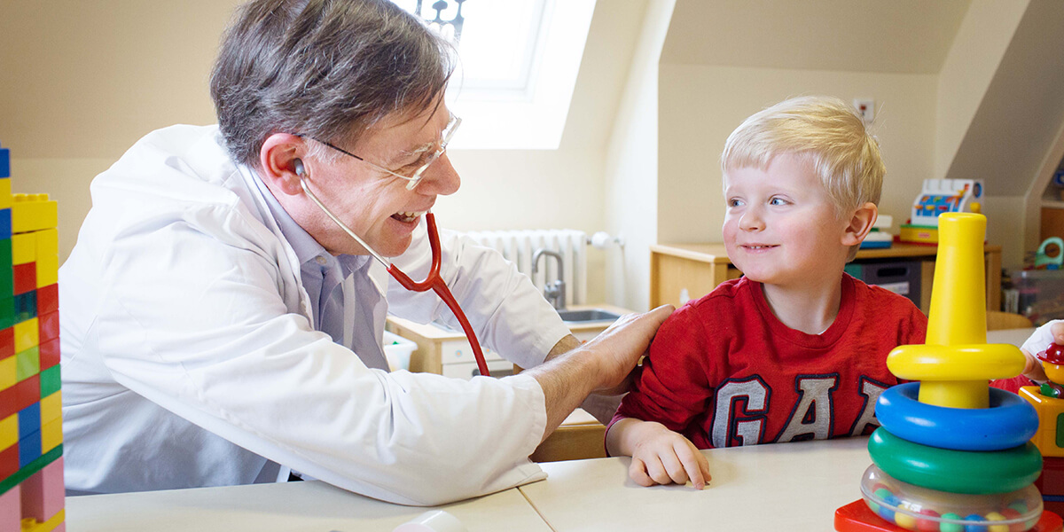 Christoph Klein trägt ein Medizinkittel und hört ein Kind, das rechts von ihm sitzt, mit einem Stethoskop ab. Beide schauen sich an und lächeln