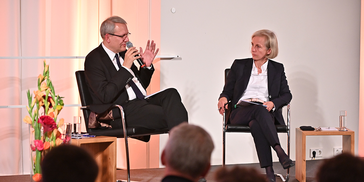 Andreas Wirsching sitzt auf einer Bühne und hält ein Mikro in der Hand. Neben ihm sitzt Frau Ursula Münch und schaut ihn an. In der Hand hält sie einen Stift und auf den Knien hat sie einen Notizblock
