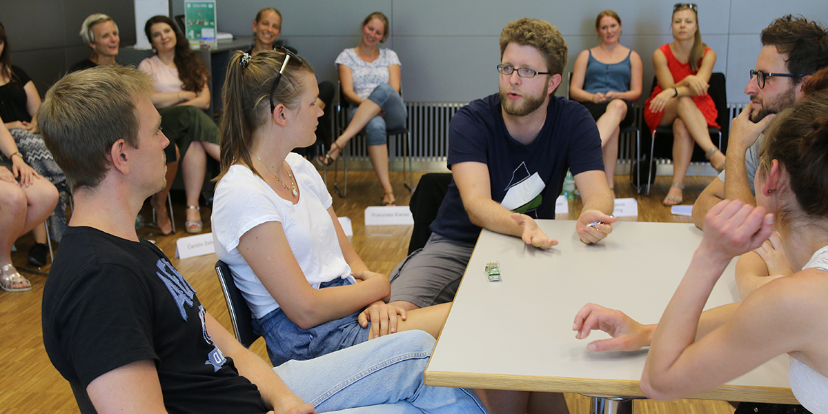 Fünf Personen sitzen an einem Tisch. Um sie herum haben mehrere Personen einen Stuhlkreis gebildet.