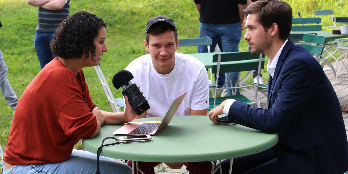 Eine Person mit weißem T-Shirt und schwarzer Cap sitzt im Garten der Akademie für Politische Bildung an einem runden Tisch und schaut direkt in die Kamera. Links von ihm sitzt eine Frau, die ein Aufnahmegerät in der Hand hält und einen Laptop sowie ein Handy neben sich liegen hat. Sie ist gerade dabei einen Mann zu interviewen, der ihr gegenüber sitzt