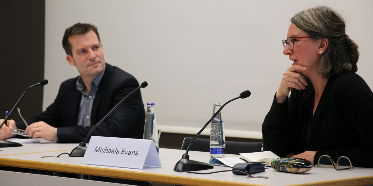 Michaela Evans sitzt an einem Tisch auf dem Podium der Akademie für Politische Bildung Tutzing und redet. Links von ihr sitzt Jörg Siegmund und hört ihr zu. Beide haben jeweils ein Mikrofon, Namensschild sowie Wasserflaschen vor sich stehen.