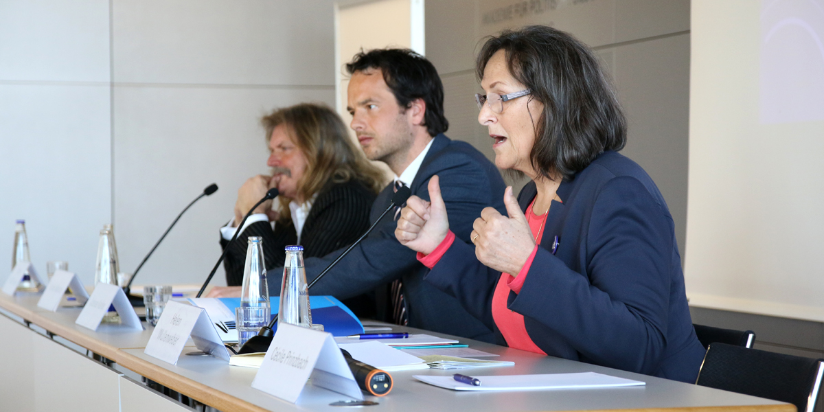 Helen Wullenweber sitzt im Hörsaal der Akademie für Politische Bildung Tutzing an einem Pult und redet. Links von ihr sitzen Andreas Kalina und Leslie Mandoki. Vor ihnen stehen jeweils ein Namensschild, Mikrofon und Wasserflaschen.