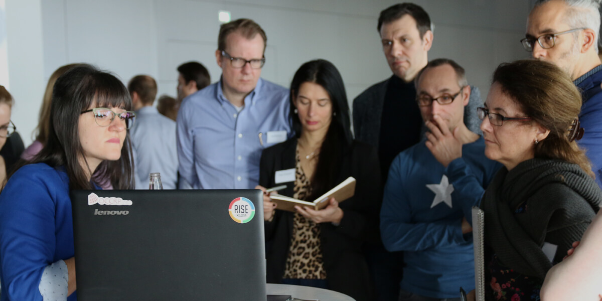 Eine Frau mit Brille und blauem Blazer steht an einem Stehtisch, auf dem ein Laptop steht und präsentiert etwas. Vor ihr stehen im Halbkreis mehrere Personen, die ihr zuhören und zum Teil Notizen machen 