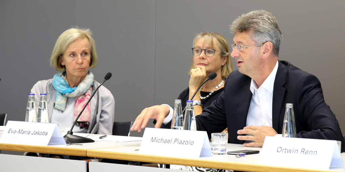 Michael Piazolo sitzt im Hörsaal der Akademie für Politische Bildung auf dem Podium an einem Tisch und redet. Links von ihm sitzen Eva-Maria Jakobs und Ursula Münch und hören ihm zu. Vor ihnen steht jeweils ein Namensschild, Mikrofon und mehrere Wasserflaschen aus Glas.