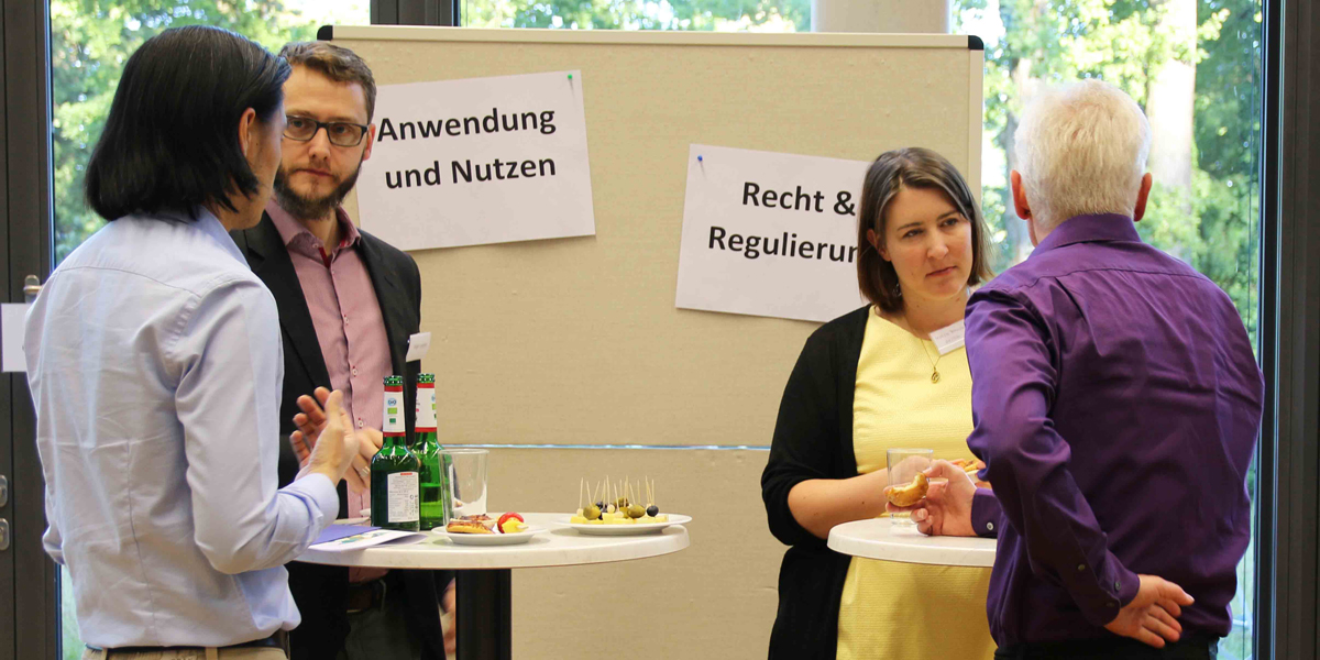 Jeweils zwei Personen stehen an einem Stehtisch mit Getränken und Essen und unterhalten sich. Im Hintergrund ist eine Pinnwand, an der zwei Schilder hängen. Auf dem einen Schild steht "Anwendung und Nutzung" und auf dem zweiten "Recht und Regulierung"