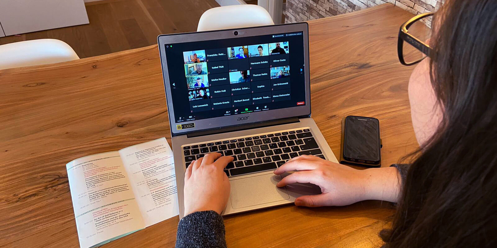 Eine Person verfolgt die Online-Tagung über einen Laptop. Links von ihr liegt das Tagungsprogramm und rechts von ihr ein Smartphone.