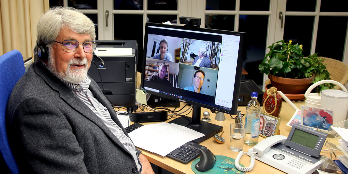 Michael Schröder sitzt am Computer in seinem Büro und lächelt in die Kamera. Im Hintergrund sind auf dem Bildschirm die Teilnehmenden des Werkstattgesprächs zu sehen. 
