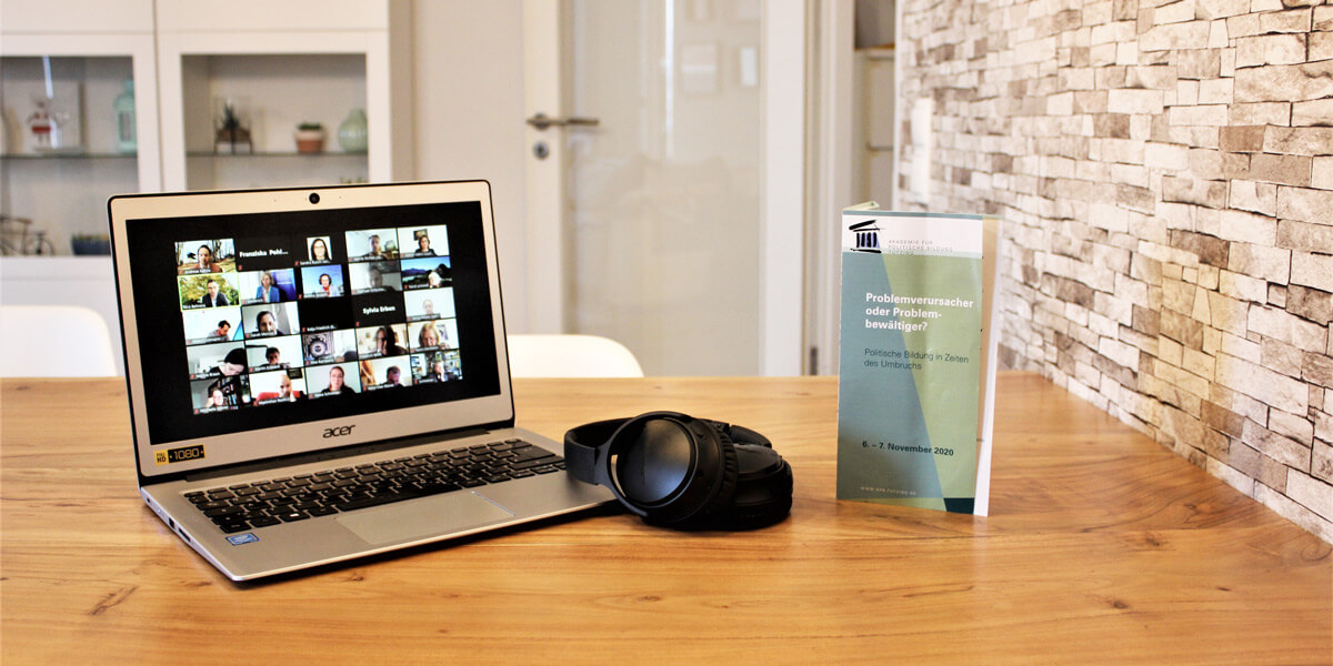 Ein Laptop, auf dem die Teilnehmenden der Online-Tagung über Zoom zu sehen sind, steht auf einem Tisch. Rechts auf der Tastatur liegen Over-Ear-Kopfhörer und daneben steht ein Flyer mit dem Tagungsprogramm.