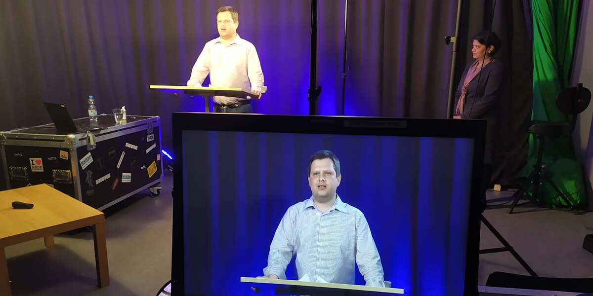  Markus Kaiser steht in einem Stuio an einem Pult und hält eine Vorlesung. Neben ihm steht Niraimathi Appavu-Gauss und schaut zu. 