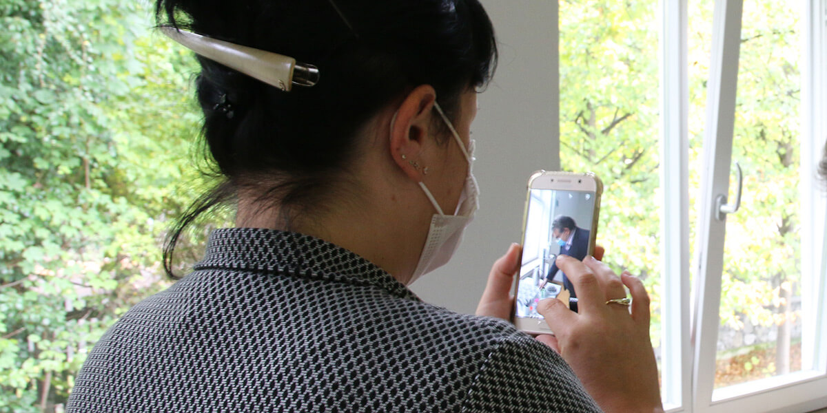 Frau mit medizinischer Maske steht mit dem Rücken zur Kamera und macht mit dem Smartphone ein Foto von einem Mann