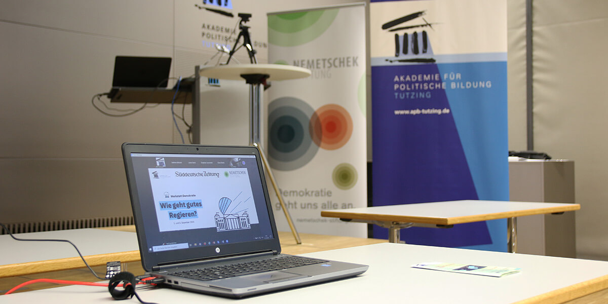 Ein Laptop steht auf einem Tisch. Auf dem Desktop ist der Titel der Werkstatt  "Wie geht gutes Regieren?" zu lesen. Im Hintergrund stehen verschiedene Roll-Ups der Akademie für Politische Bildung Tutzing sowie ein Stehtisch und ein weiterer Laptop