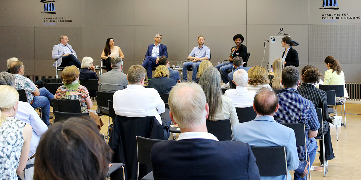 Zu sehen ist ein Podium auf dem eine Referentin und fünf Referenten im Halbkreis sitzen. Davor steht eine Moderatorin. Im Vordergrund sind die Hinterköpfe des Publikums zu sehen.