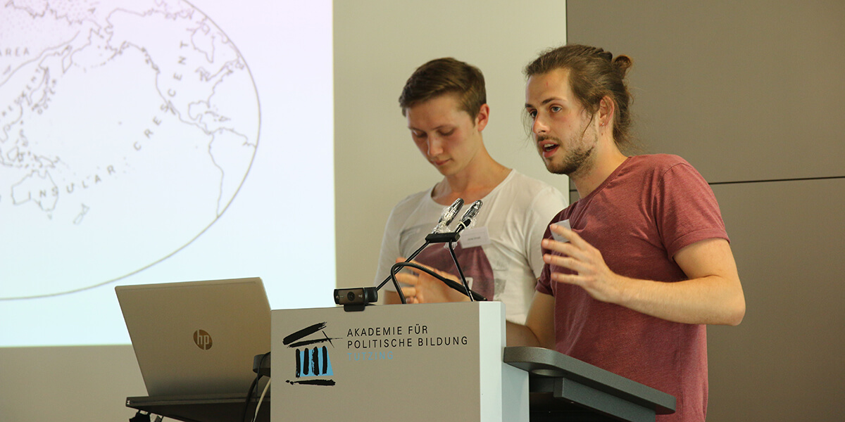 Jonas Arnold (links) und Jonathan Icks (rechts), Studierende der Philipps-Universität Marburg, stehen nebeneiander auf dem Podium der Akademie für Politische Bildung und tragen vor. Direkt vor ihnen steht ein Laptop, im Hintergrund wird eine Power-Point-Präsentation vom Beamer an die Wand geworfen.