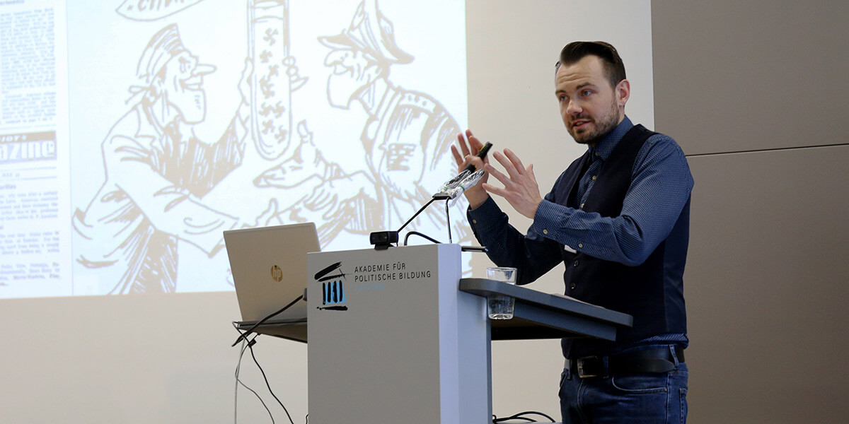 Christopher Nehring steht am Pult der Akademie für Politische Bildung, gestikuliert und spricht in ein Mikrofon. Hinter ihm ist eine Präsentation mit einer Karikatur zu sehen.