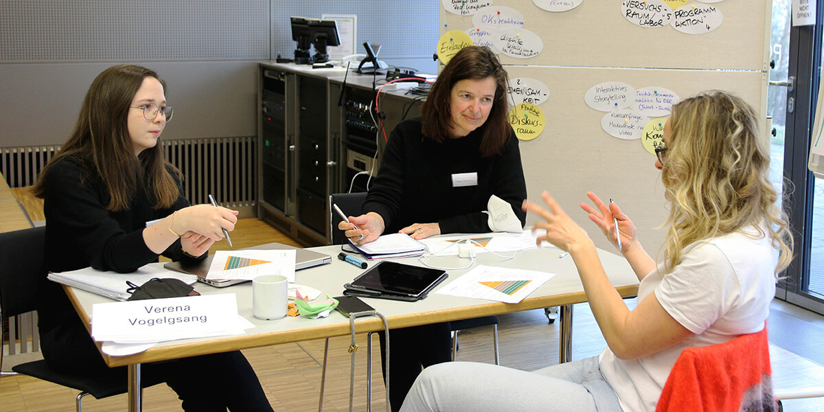 Drei Frauen sitzen um einen Tisch mit Tablet, Papier und Stiften und arbeiten in der Gruppe