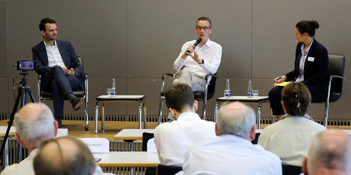 Auf dem Podium der Akademie für Politische Bildung spricht Michael Böhmer in ein Mikro. Links von ihm sitzt Florian Dorn und rechts von ihm Giulia Mennillo. Im Vordergrund sind Köpfe des Publikums zu sehen.