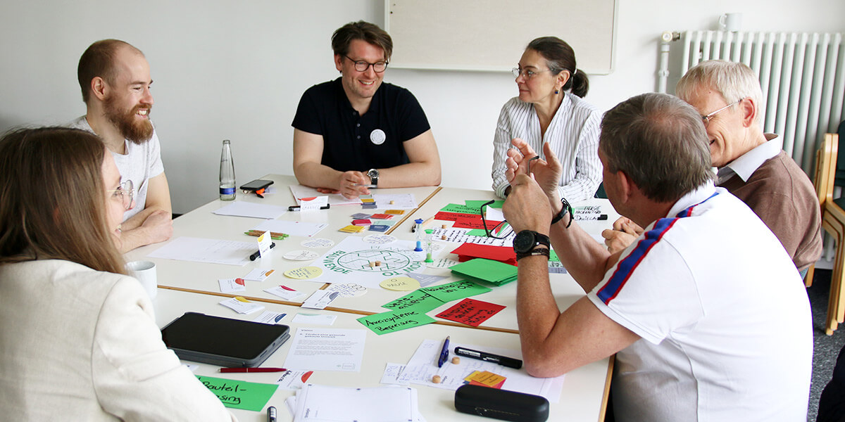 Eine Gruppe von sechs Menschen sitzt um einen Tisch, auf dem Filzstifte und bunte Kärtchen reden, lacht und diskutiert.