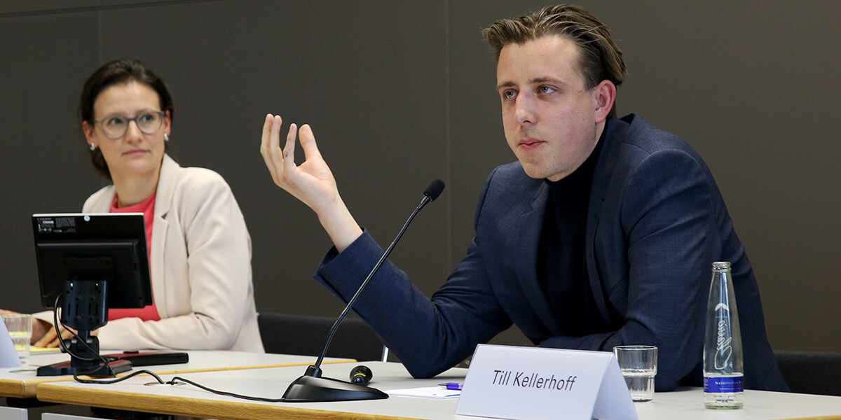 Giulia Mennillo und Till Kellerhoff sitzen an der Akademie für Politische Bildung auf dem Podium. Tim Kellerhoff spricht und Giulia Mennillo wendet sich ihn zu.