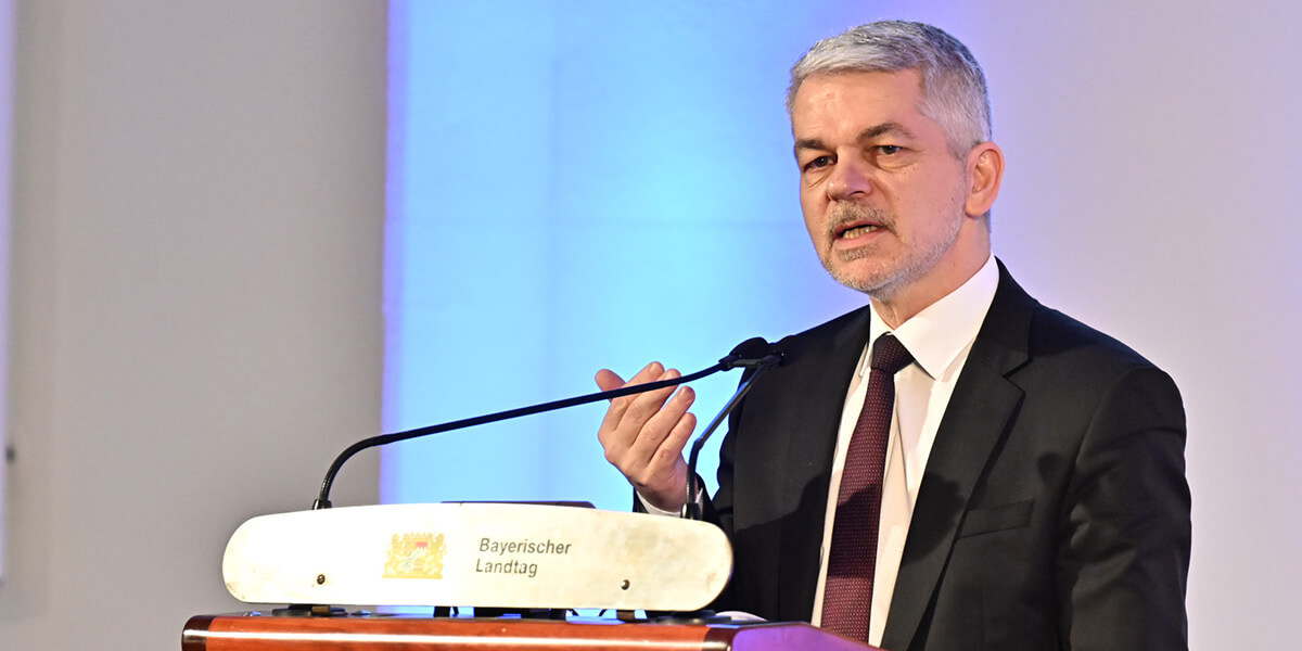 Carlo Masala steht an einem Podium mit der Aufschrift "Bayerischer Landtag" und spricht.