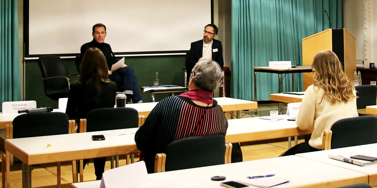 Cengiz Günay sitzt links auf dem Podium der Akademie für Politische Bildung und spricht ins Publikum, rechts neben im sitzt Hakan Akbulut. Im Vordergrund sind Tische und Menschen im Publikum von hinten zu sehen.