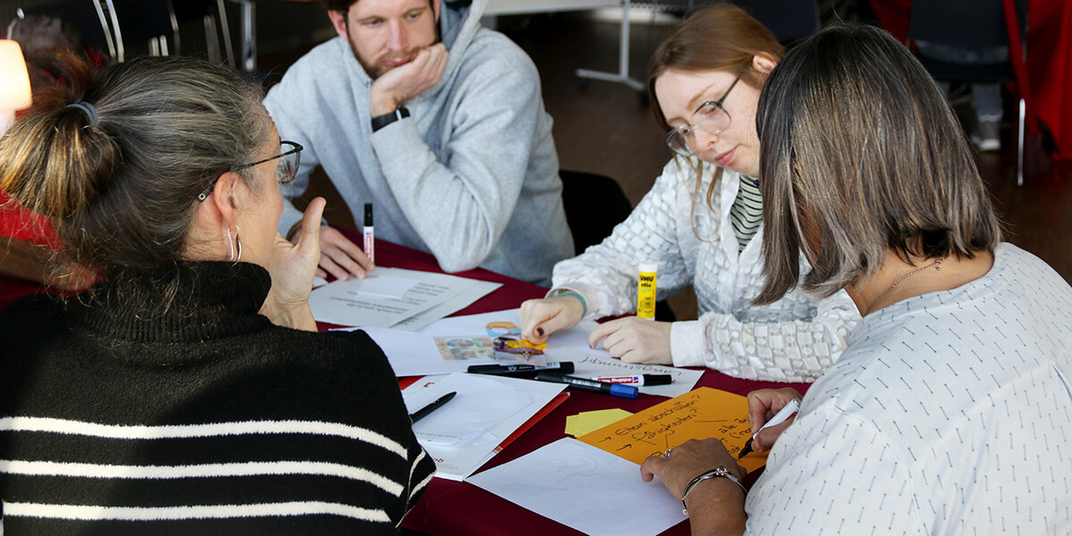 Vier Teilnehmerinnen und Teilnehmer sitzen um einen Tisch und arbeiten gemeinsam. Zwischen ihnen liegen bunte Blätter und Stifte verteilt.