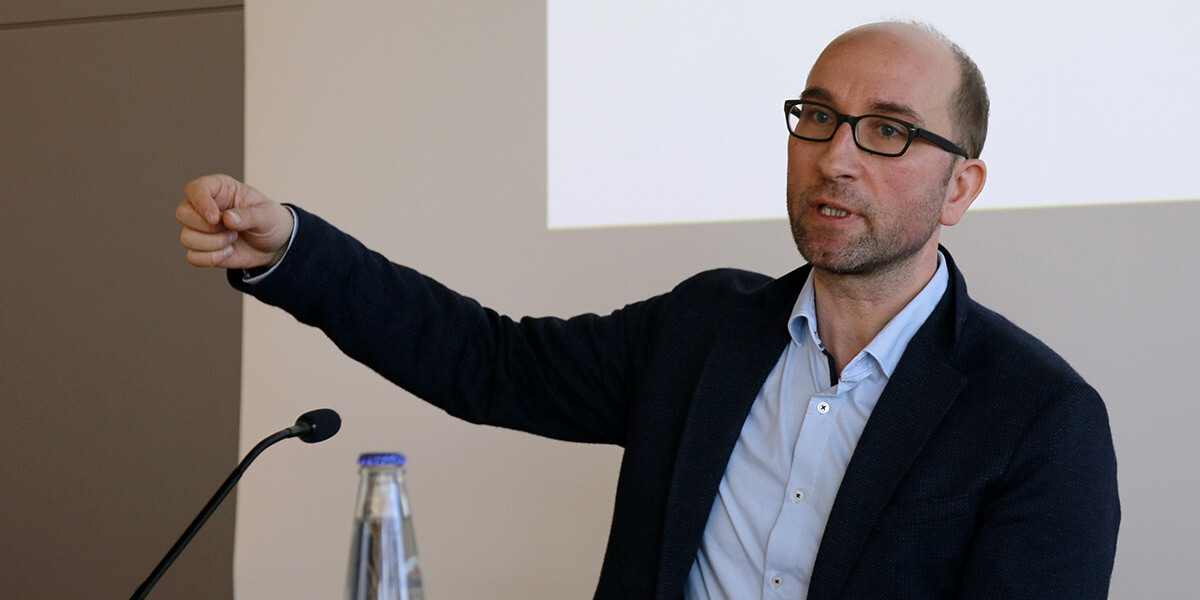 Magnus Treiber sitzt auf dem Podium der Akademie für Politische Bildung, hinter ihm eine Leinwand. Er spricht und gestikuliert.