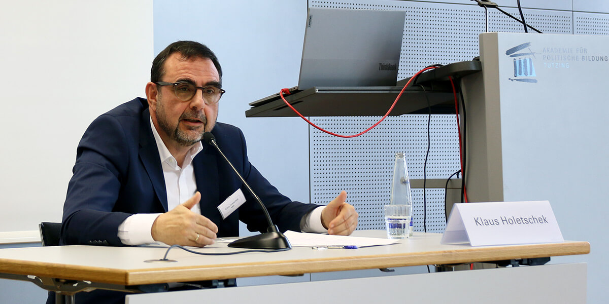 Klaus Holetschek sitzt auf dem Podium der Akademie für Politische Bildung, spricht und gestikuliert. Hinter ihm ist ein Pult mit Laptop zu sehen.