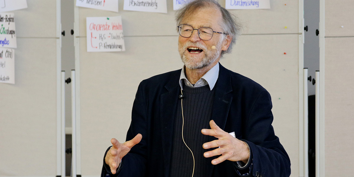 Nikolaus Geiler steht vor mehreren Stellwänden mit bunt beschriebenen Zetteln, gestikuliert und spricht in ein Headset.