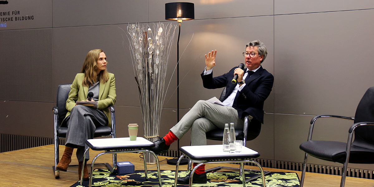 Benedikt Franke sitzt auf dem Podium der Aakdemie und spricht in ein Mikrofon. Links von ihm sitzt Anja Opitz und sieht ihn an.