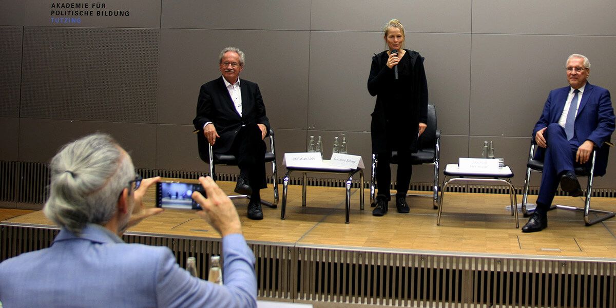 Christian Ude, Dorothee Schiwy und Joachim Herrmann sind am Podium. Dorothee Schiwy spricht ins Mikrofon und Michael Mayer fotografiert mit seinem Handy das Podium aus dem Publikum.