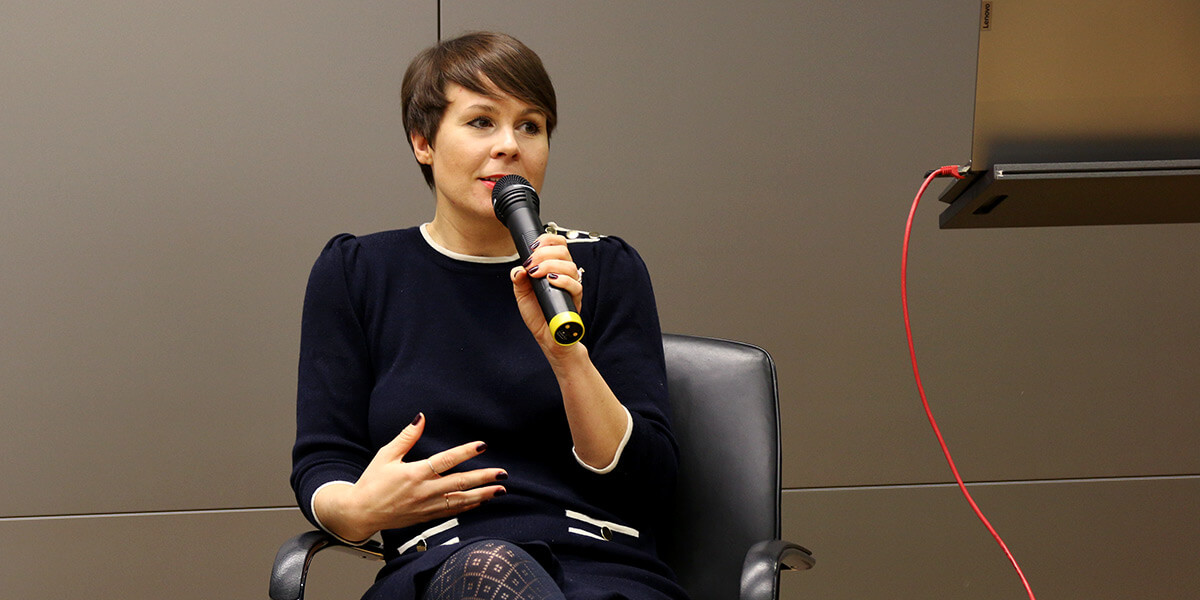 Elisabeth Hoffberger-Pippan sitzt auf dem Podium der Akademie für Politische Bildung. In der linken Hand hält sie ein Mikrofon, in das sie spricht. Mit der rechten Hand gestikuliert sie.