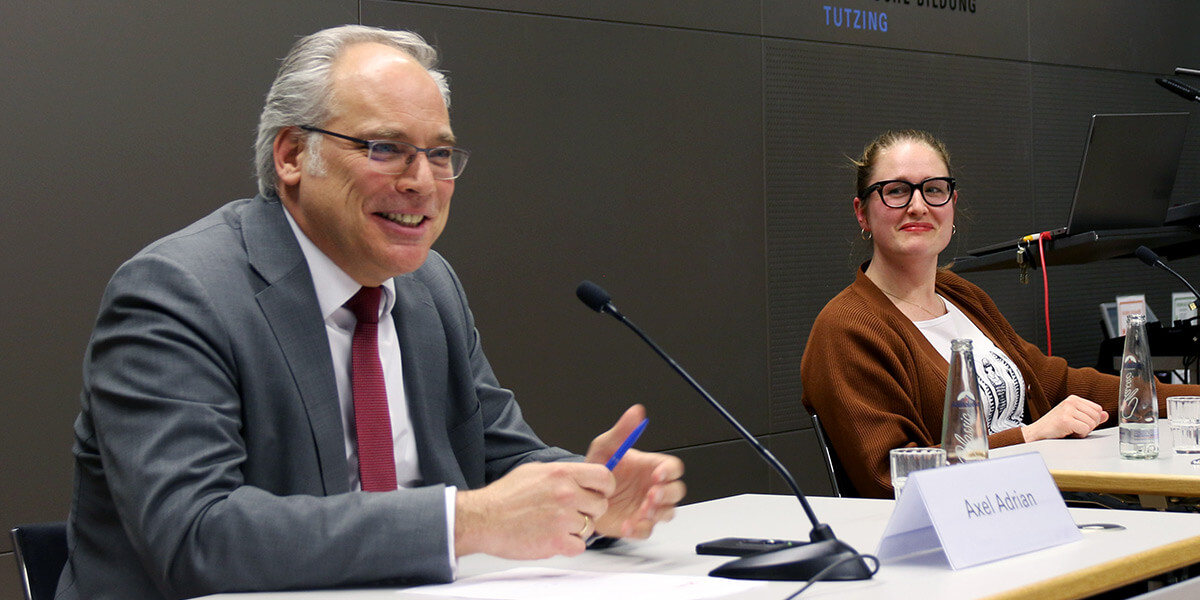 Axel Adrian sitzt auf dem Podium der Akademie für Politische Bildung und spricht in ein Mikrofon. Rechts sitzt Julia Post und wendet sich ihm zu.