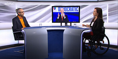 Links sitzt Michael Mayer an einem halb runden Tisch im Studio von alpha-demokratie, rechts Mirjam Kottmann. Im Hintergrund ist ein Bildschirm zu sehen