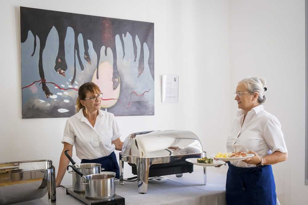 Renate Brunner steht an der Essensausgabe der Akademie für Politische Bildung und spricht mit Jenny Weber, die Teller trägt