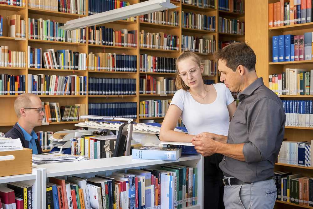 Links sitzt Moritz Fink an einem Schreibtisch in der Bibliothek der Akademie für Politische Bildung. Rechts steht Thomas Schölderle und spricht mit einer Besucherin mit Blick in ein Buch.