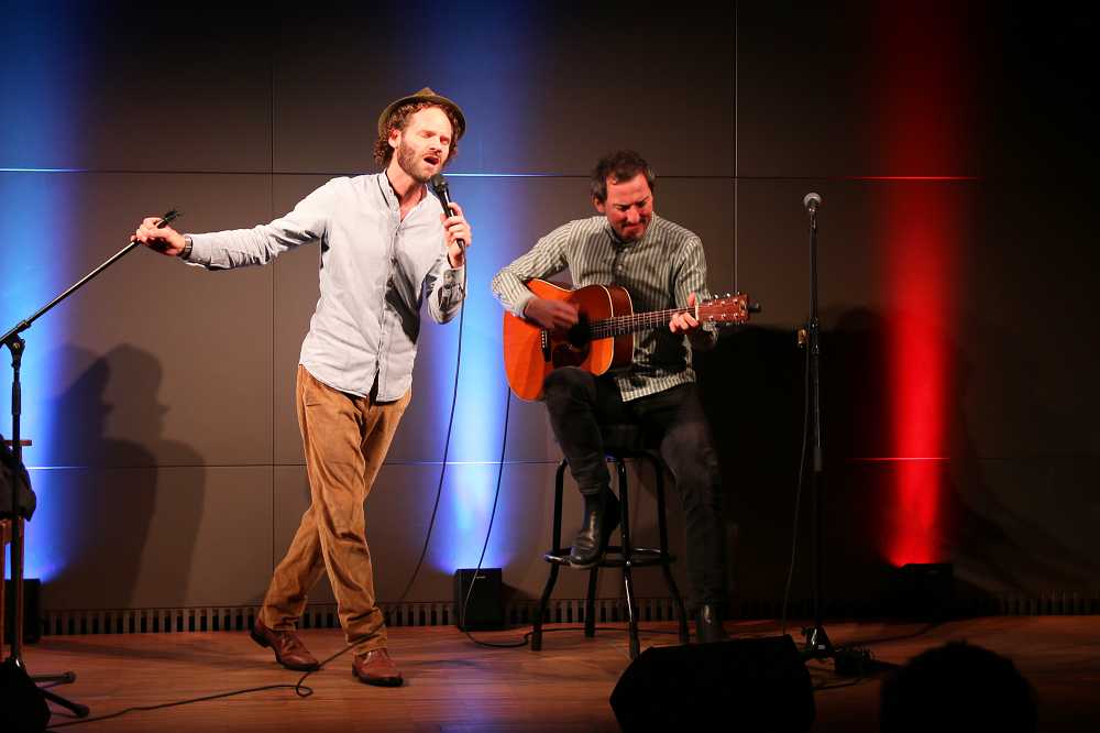 Maxi Schafroth hält mit der rechten Hand den Mikrofonständer, in der linken ein Mikrofon, singt und geht beim Tanzen auf der Bühne der Akademie für Politische Bildung in die Knie. Neben ihm sitzt Markus Schalk auf einem Barhocker und spielt Gitarre.