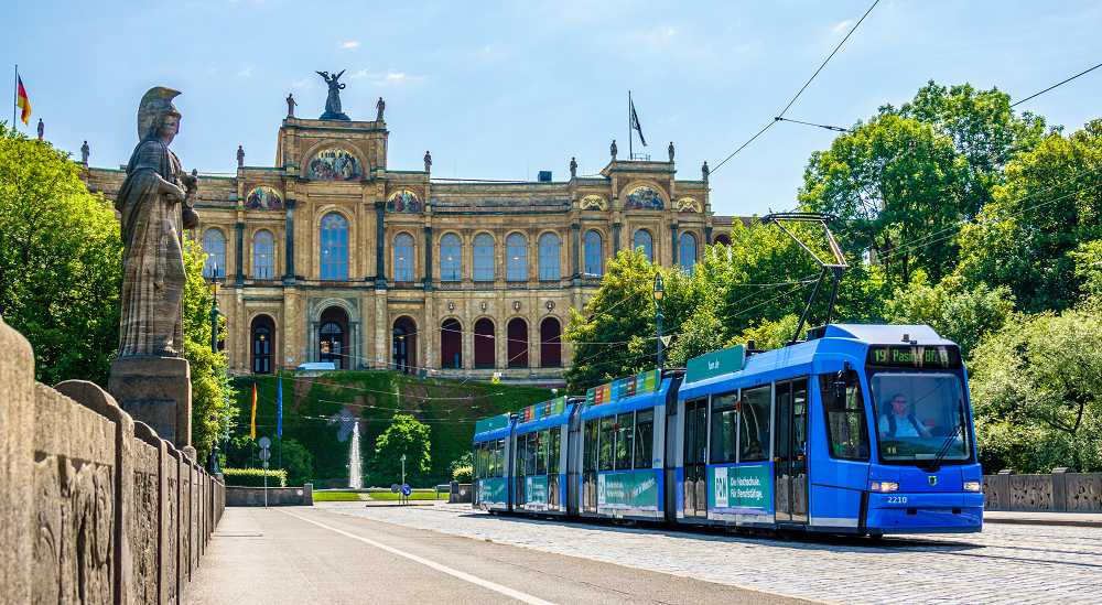 Eine blaue Trambahn fährt vom Maximilianeum in München weg. Im Hintergrund sind Bäume und blauer Himmel.