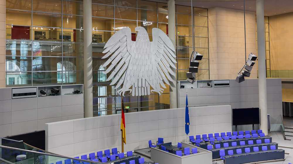 Der Plenarsaal des Deutschen Bundestags mit dem silbernen Adler an der Glaswand und blauen Sitzen.